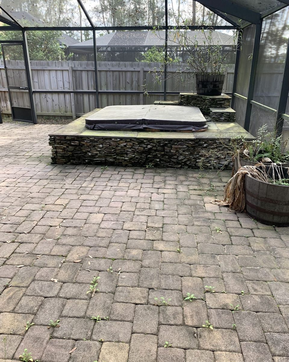 A screened lanai with gray pavers and weeds around the hot tub