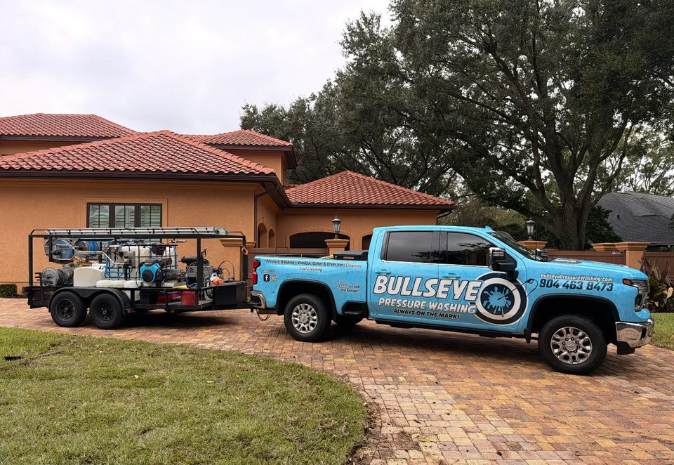 The light blue Bullseye Pressure Washing pickup and its equipment trailer on a paver driveway in front of a stucco home with a tile roof